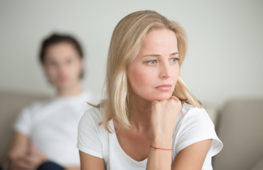 Eine Frau ist nachdenklich und verzweifelt, im Hintergrund sitzt Ihr Mann - Unterstützung Paartherapie bei Affäre.
